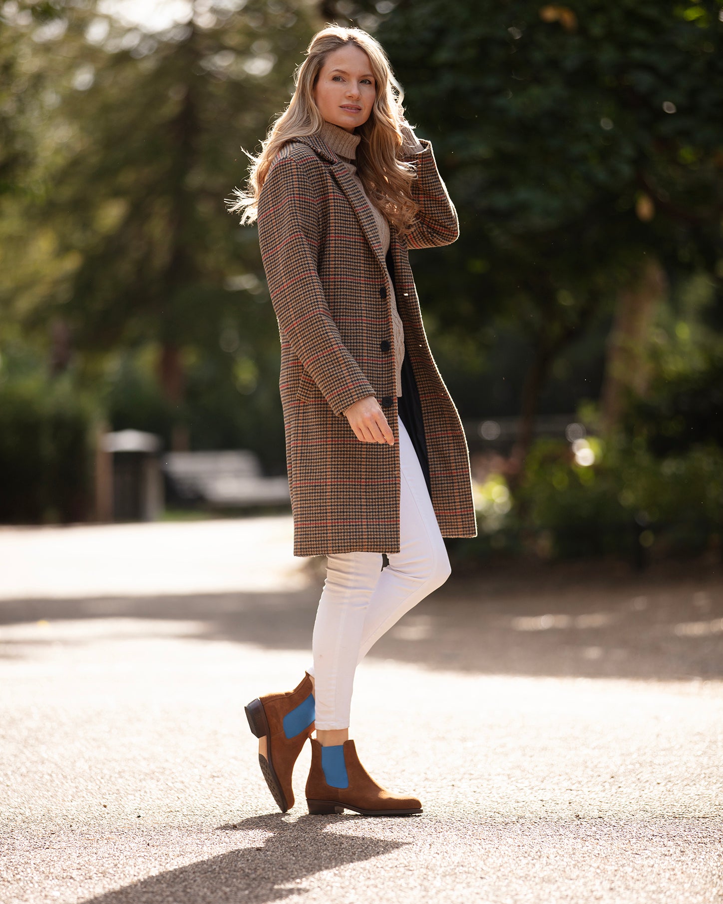 Woman wearing a brown checkered coat and white pants walking outdoors.