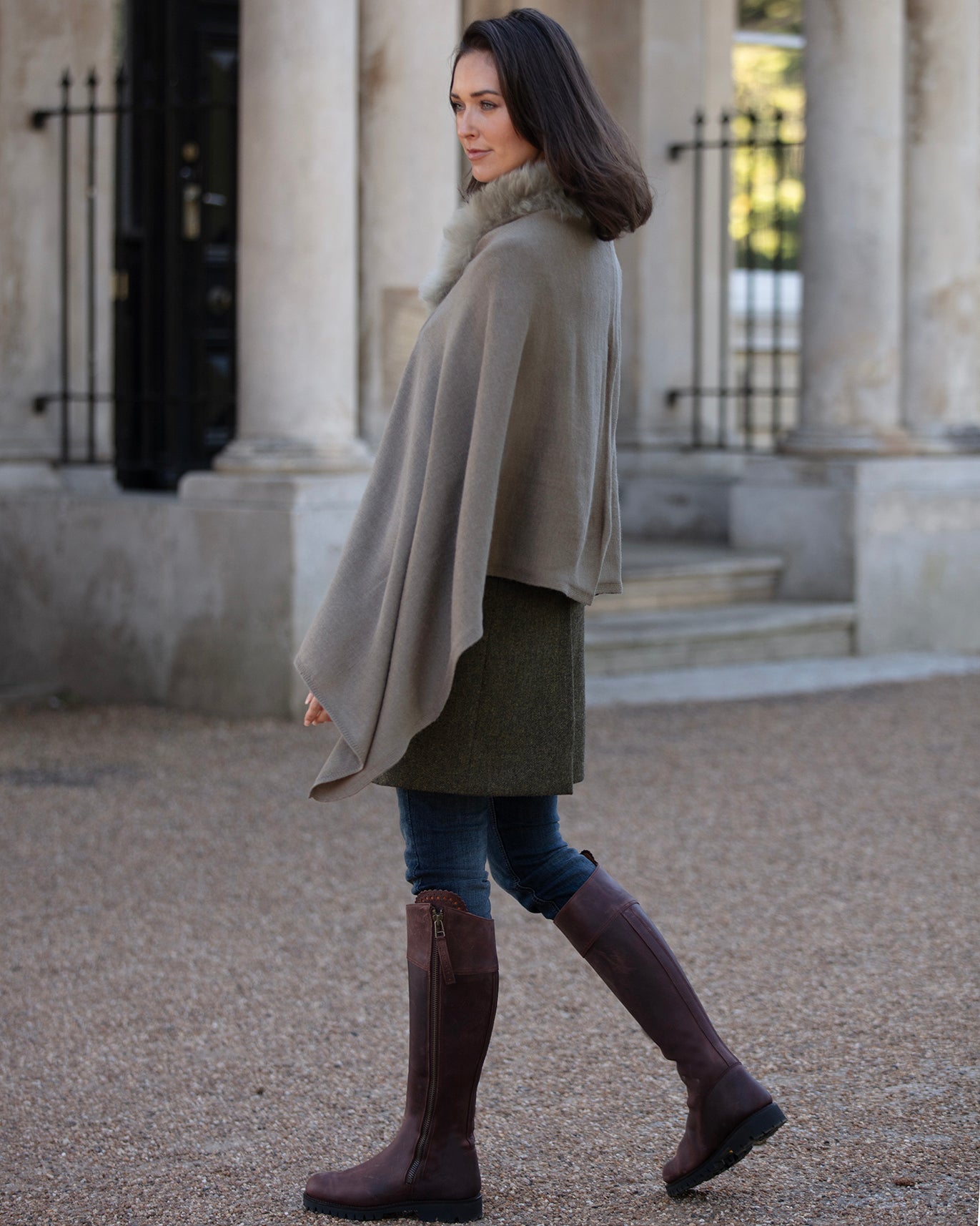 Woman wearing a beige cape and brown knee-high boots walking outdoors.