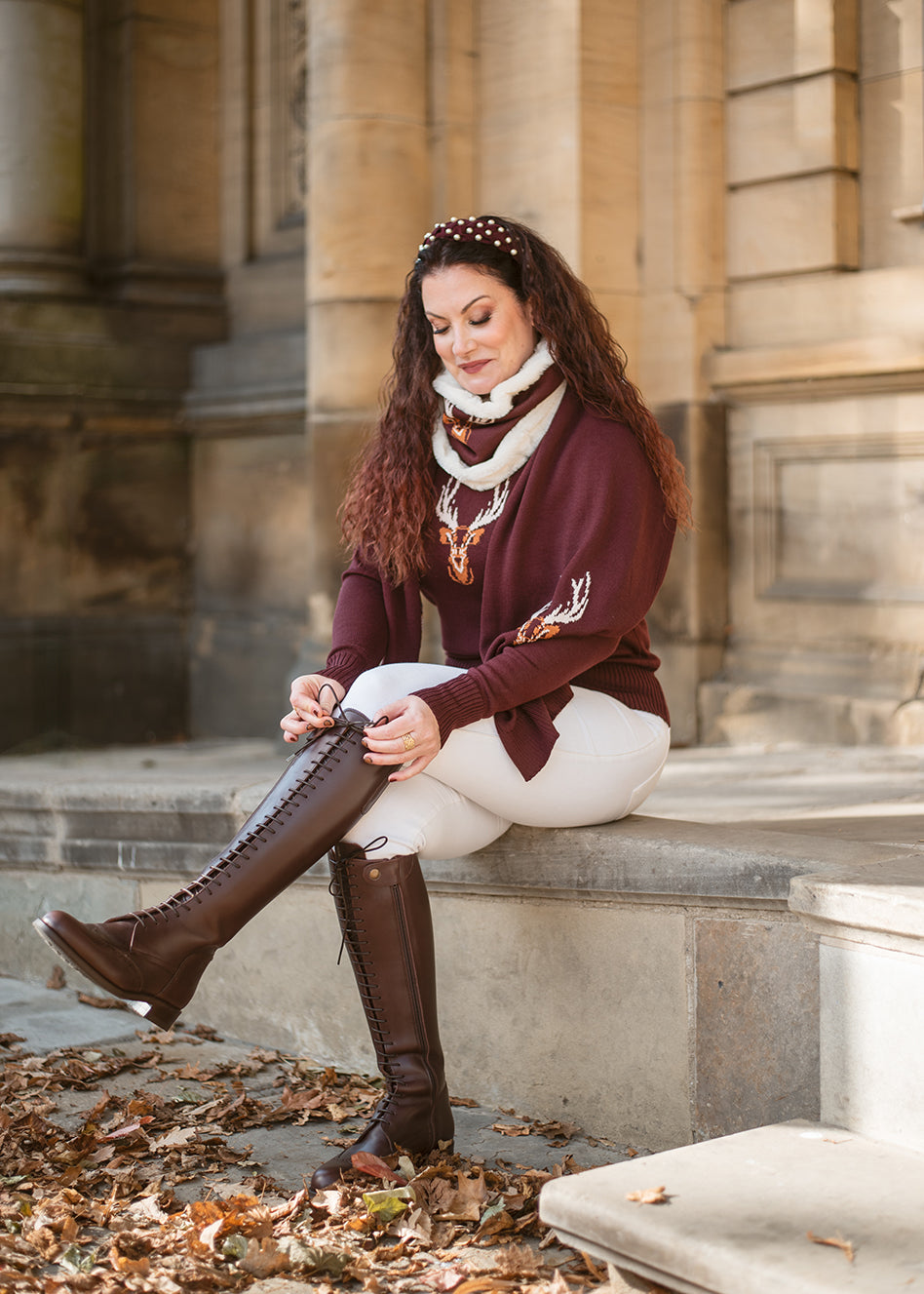 Woman sitting on steps wearing a burgundy sweater, white pants, and knee-high brown boots.