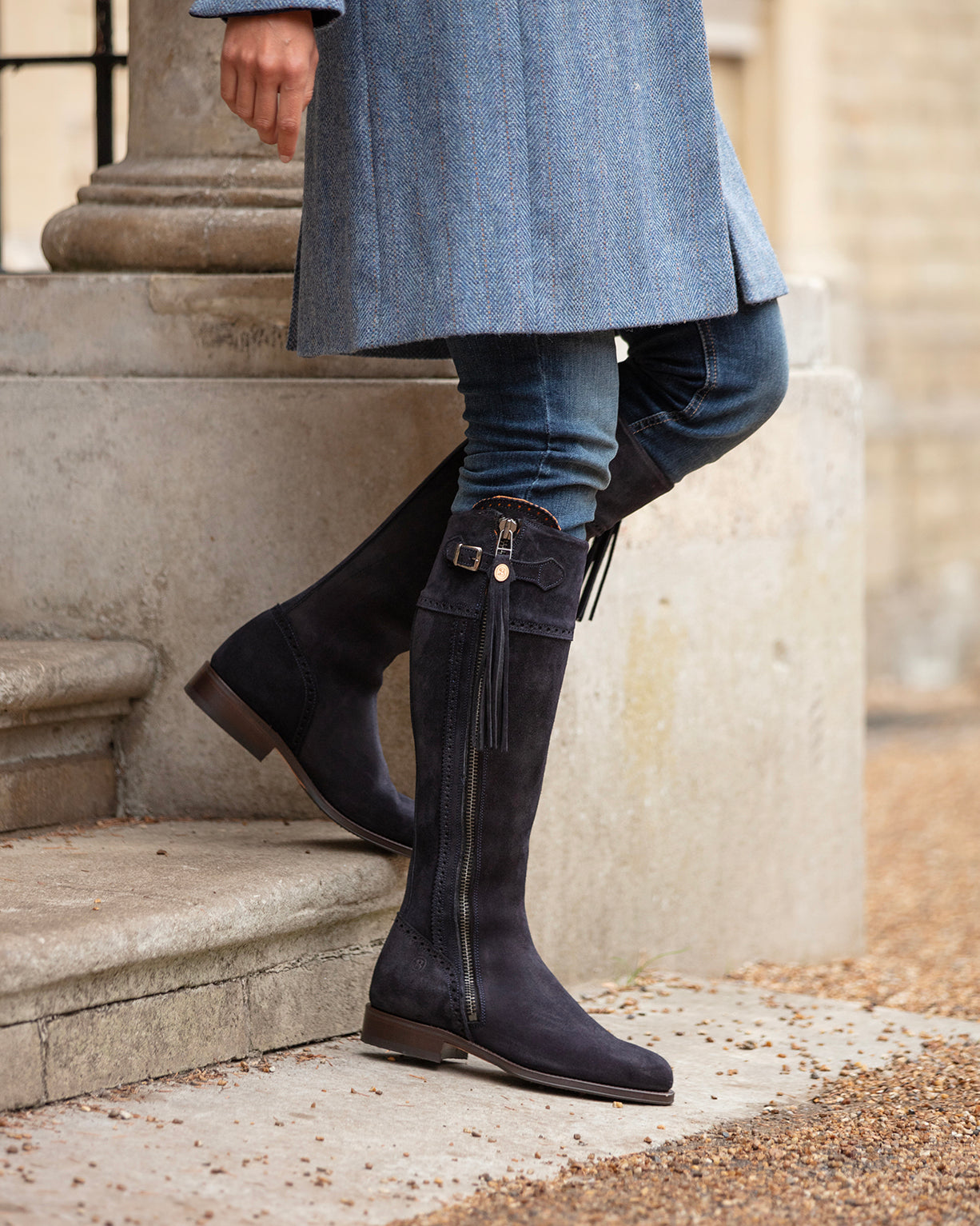 Person wearing navy suede knee-high boots and a blue coat on stone steps.