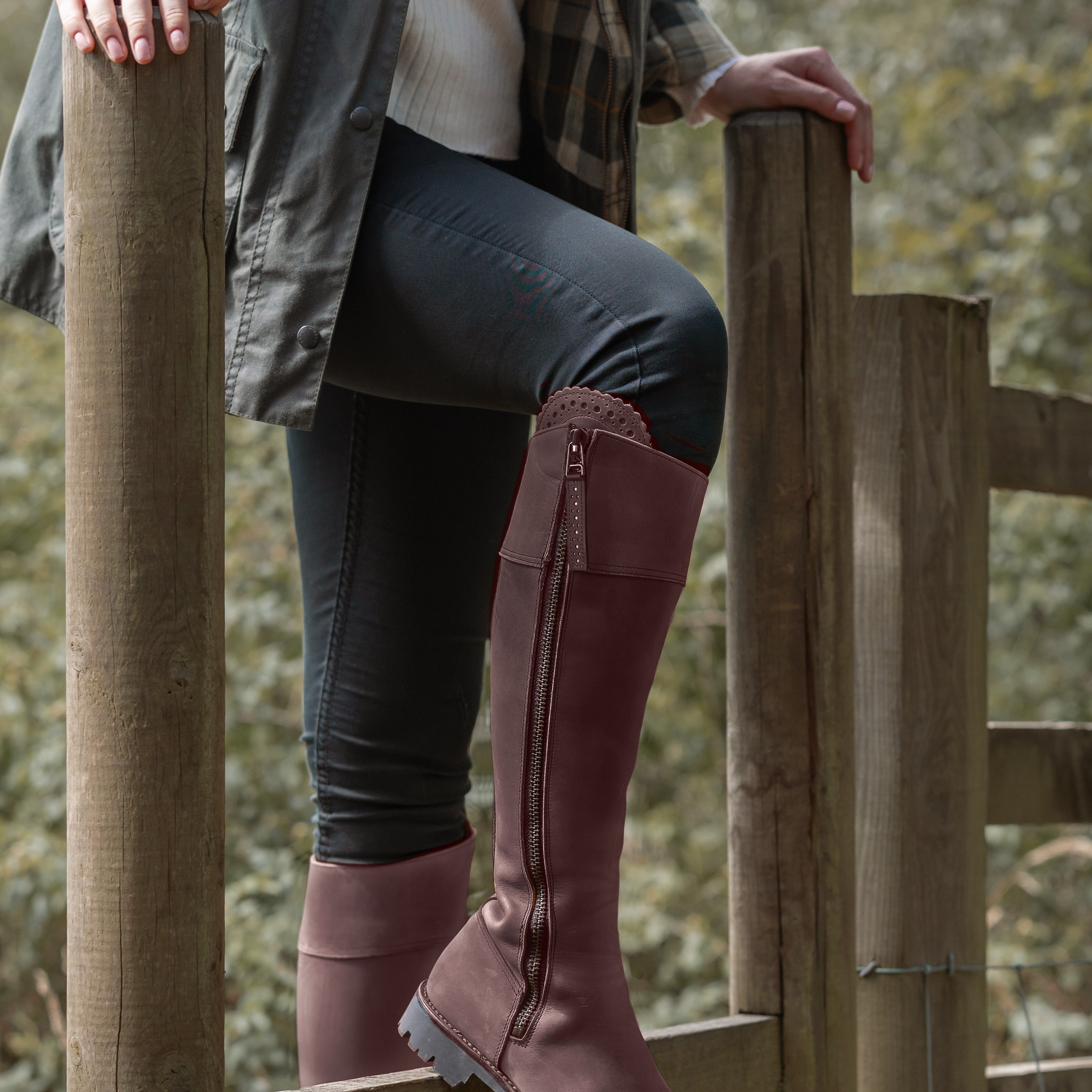 Person wearing brown knee-high boots standing on a wooden fence with a natural background.